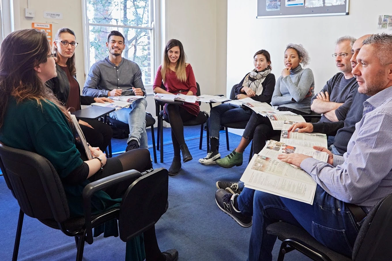 A group of diverse adults is listening to the teacher in a small classroom.